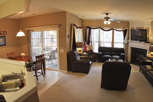 A cozy living room and dining area in a residential facility. The living room features dark leather sofas and armchairs arranged around a fireplace with a flat-screen TV mounted above it. Large windows with curtains allow natural light to fill the room. Adjacent to the living room is a dining area with a wooden table and chairs, next to sliding glass doors that lead to an outdoor patio with additional seating. The kitchen sink and counter are partially visible in the foreground.