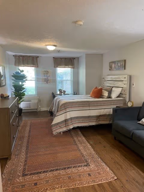 A cozy bedroom with a wooden bed frame, striped bedding, and several pillows including an orange one. There is a dark gray armchair to the right, a wooden dresser with a plant and decorative items on the left, and two windows with valance curtains at the far end. A large patterned rug covers the wooden floor, and a small table with two chairs is positioned near the windows. The walls are light-colored with framed artwork.