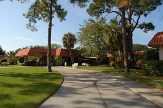 A paved driveway leading through a landscaped outdoor area with green grass, tall trees, and bushes. There are buildings with red-tiled roofs on either side of the driveway, and white chairs are visible near the center of the image under the shade of trees.
