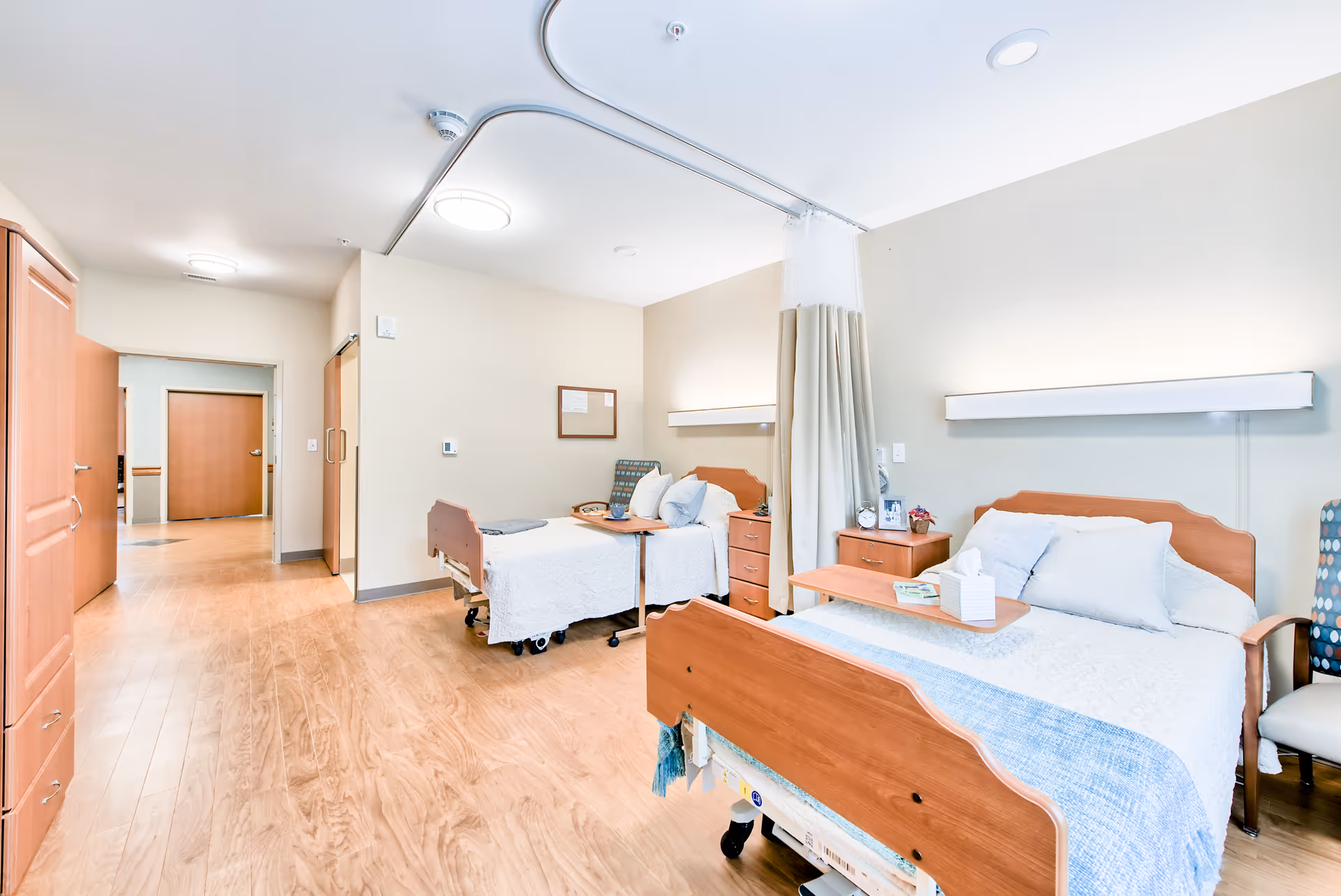 A bright and clean room in a senior living facility with two hospital-style beds separated by a privacy curtain. Each bed has white bedding and a wooden headboard. There are bedside tables with lamps, a chair, and a wooden floor. The walls are painted light beige, and the room has ceiling lights.