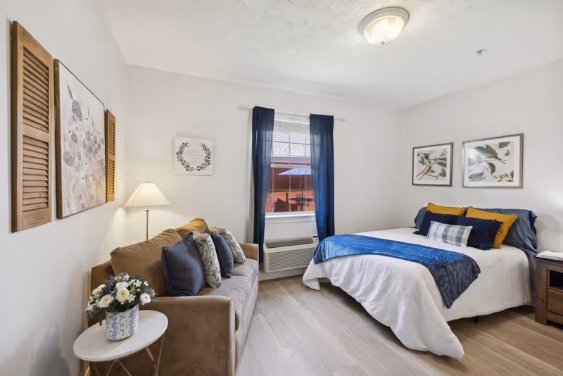 A cozy bedroom with a bed dressed in white and blue bedding, accented with yellow and plaid pillows. To the left of the bed is a brown sofa with multiple cushions, a side table with a floral arrangement, and a floor lamp. The room has light-colored walls and flooring, a window with dark blue curtains, and framed artwork on the walls.