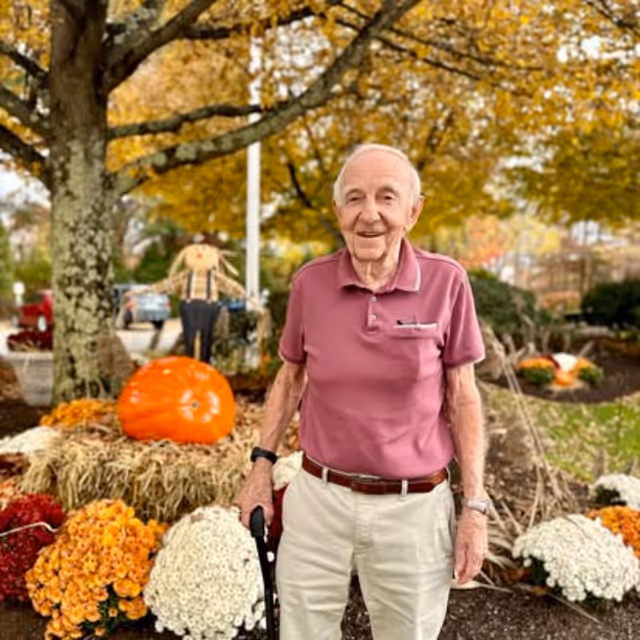 An elderly man wearing a maroon polo shirt and beige pants stands outdoors in a garden area decorated with autumn flowers, a large orange pumpkin, and a scarecrow. Trees with yellow leaves are visible in the background.