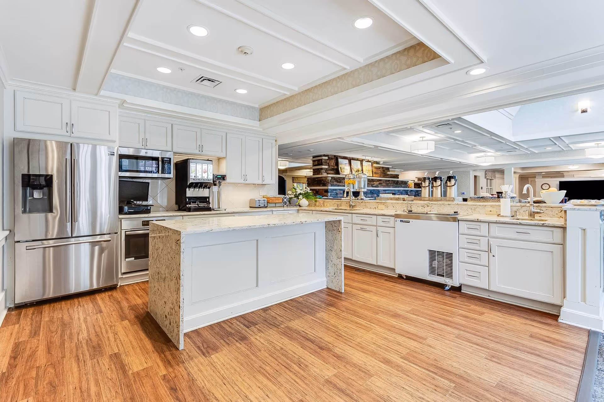 A spacious, modern kitchen area with white cabinetry, a large central island, stainless steel refrigerator, microwave, and oven. The kitchen features granite countertops, a coffee machine, and multiple sinks. The flooring is wood, and the ceiling has recessed lighting.