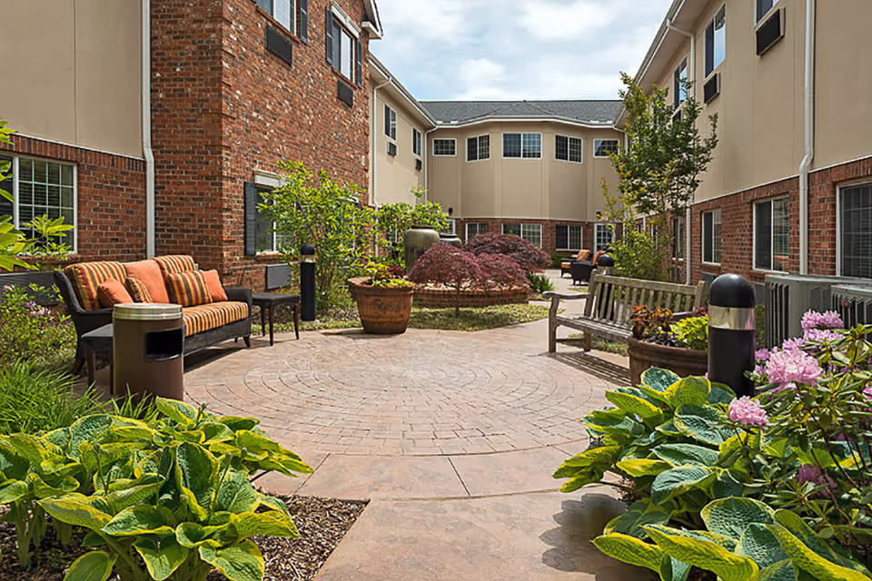 Outdoor courtyard area at Lake Pointe Landing Retirement Village featuring brick and beige building walls, a circular paved patio, benches with cushions, potted plants, green shrubs, and small trees under a partly cloudy sky.