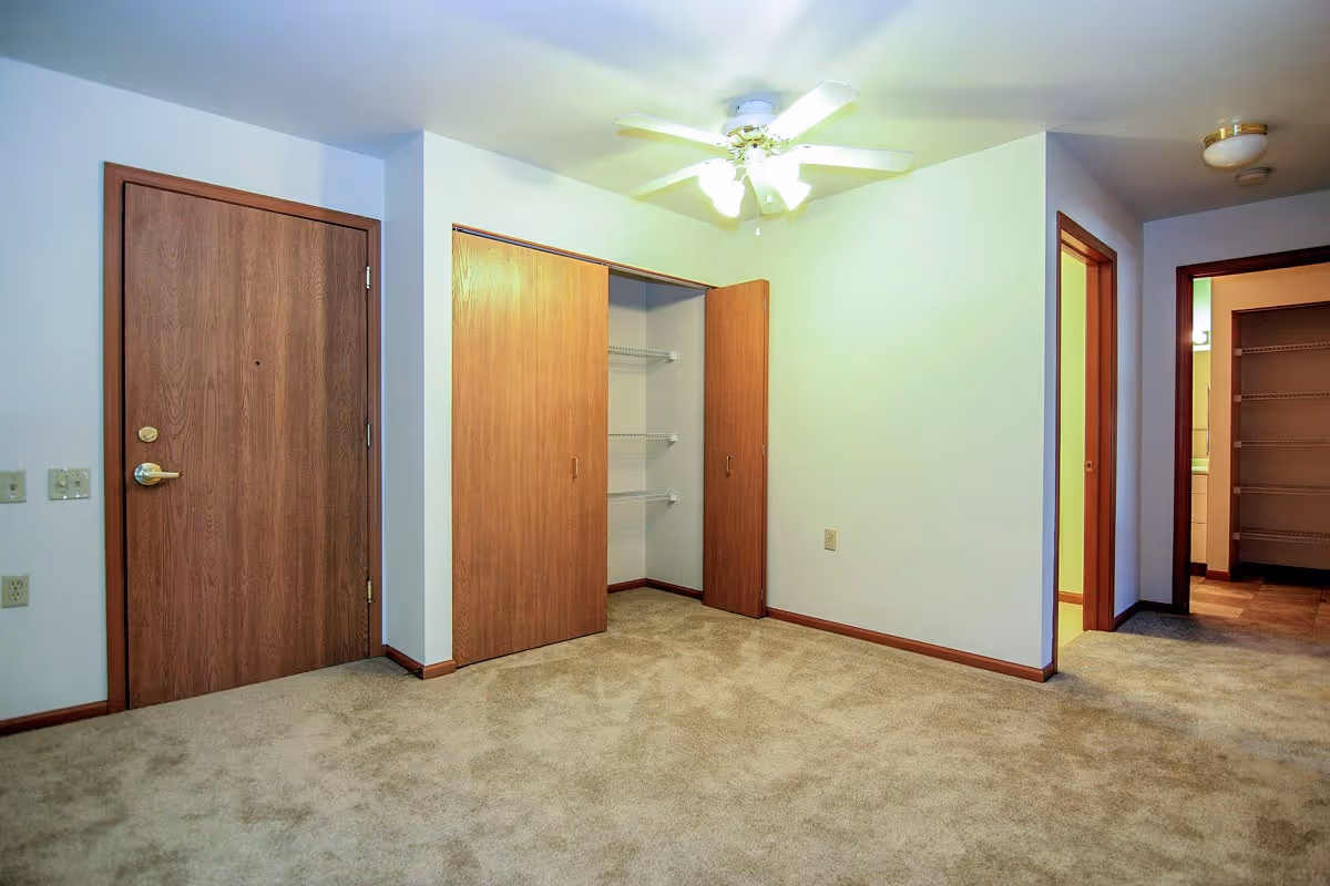 Empty room with beige carpet, white walls, a wooden door, an open closet with wooden sliding doors, and a ceiling fan with lights. There are two doorways leading to other rooms, one showing a glimpse of a kitchen area.