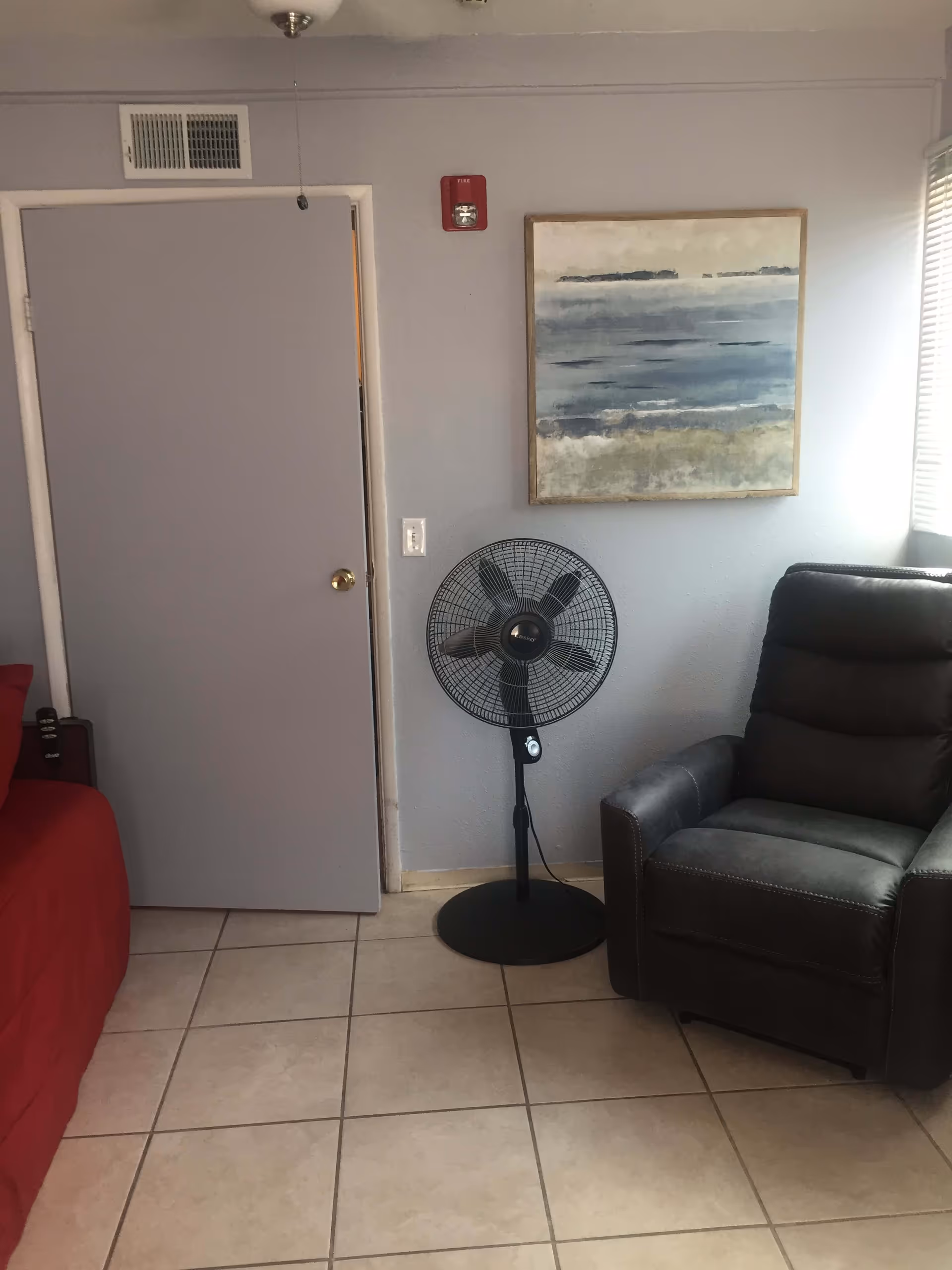 A corner of a room with a closed gray door, a standing black fan, a dark gray armchair, and a red bed partially visible. There is a painting with abstract blue and beige tones hanging on the light gray wall above the fan. The floor is tiled with beige tiles, and sunlight is coming through a window with blinds on the right side.