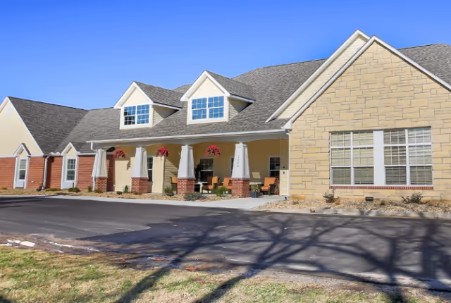 Exterior view of a single-story assisted living facility building with beige siding, stone accents, and a covered porch with columns. The porch has hanging flower baskets and outdoor seating. The sky is clear and blue.