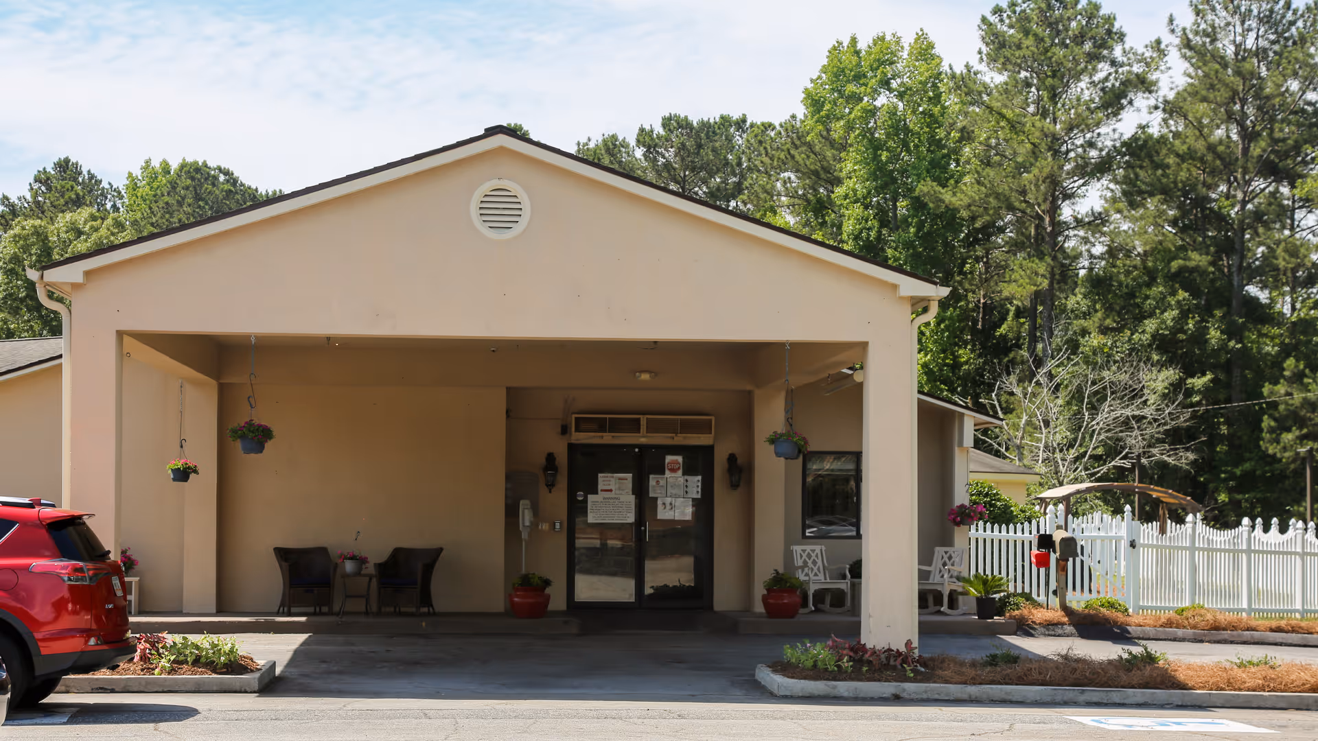 Front entrance of a beige building with a covered driveway. There are two chairs and a small table with potted plants under the covered area. Hanging flower pots are suspended from the ceiling. A red car is parked on the left side, and a white picket fence with greenery is visible on the right side.