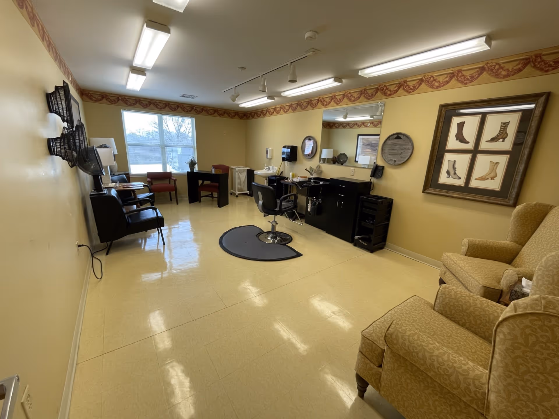 A well-lit room with beige walls and tiled floor, featuring salon chairs and hair drying stations on the left, a central styling chair with a mirror and counter on the right, and comfortable armchairs in the foreground. The room has a window letting in natural light and decorative framed pictures on the walls.