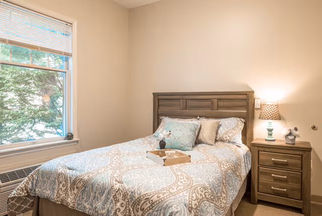 A cozy bedroom with a wooden bed frame and a bed made with a patterned blue and beige comforter and multiple pillows. There is a wooden nightstand next to the bed with a lamp and a small decorative item. A window with blinds is on the left side, letting in natural light and showing greenery outside.