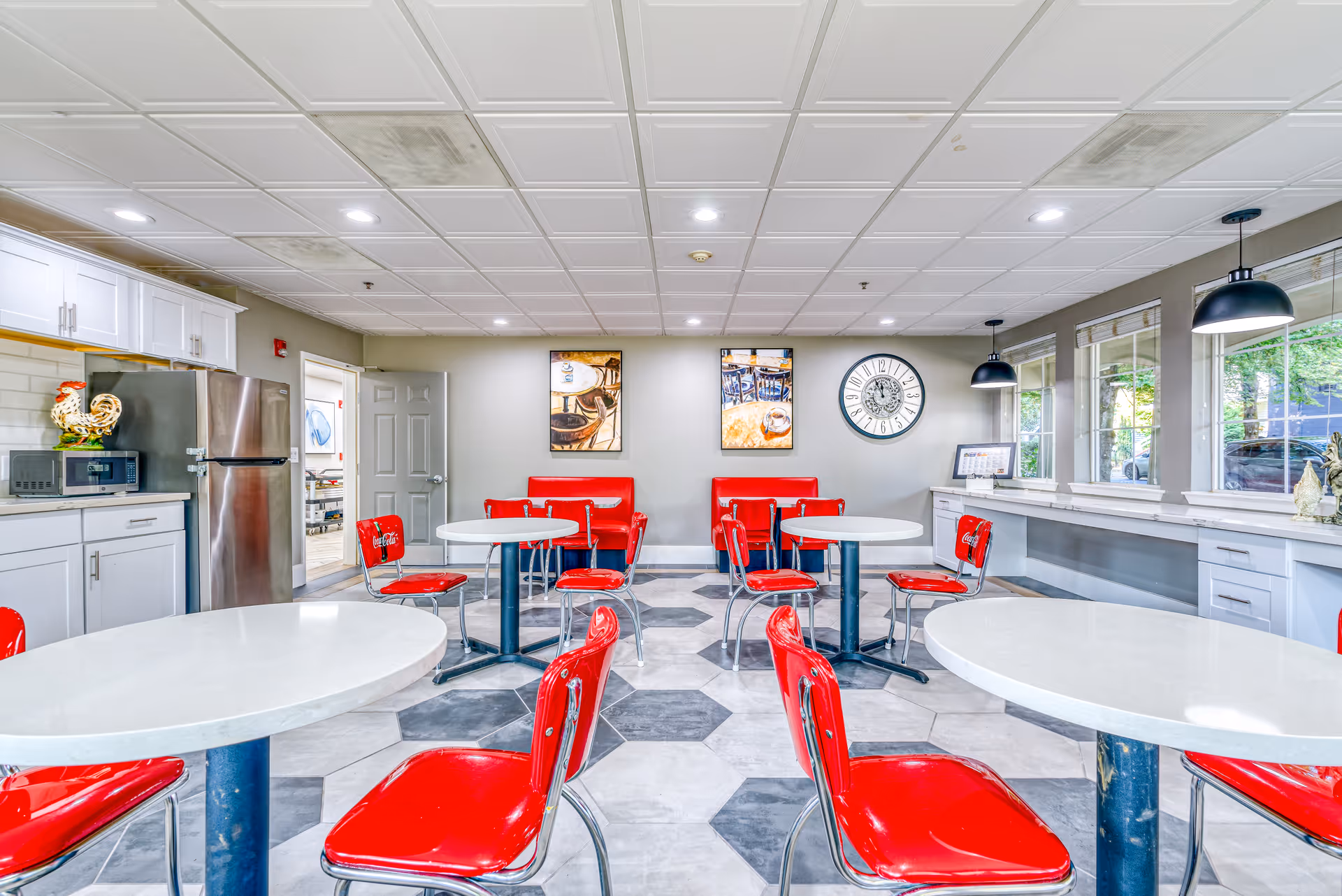 Bright dining room with white round tables and red retro-style chairs. The room features a tiled floor with hexagonal patterns, white cabinetry with a microwave and stainless steel refrigerator on the left, and large windows on the right letting in natural light. There are two red cushioned bench seats against the far wall, two framed pictures, and a large clock above them. Pendant lights hang over a counter beneath the windows.