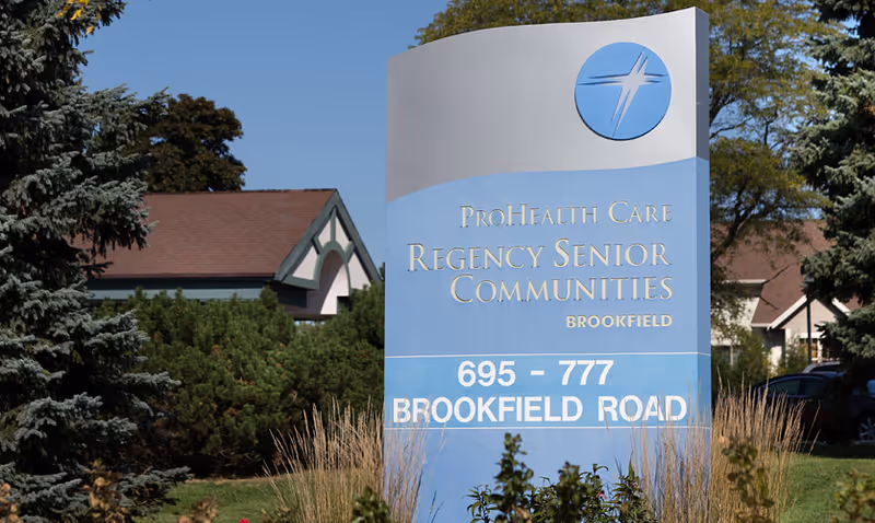 Outdoor blue sign for ProHealth Regency Senior Communities Brookfield showing the address 695–777 Brookfield Road in a landscaped front yard.