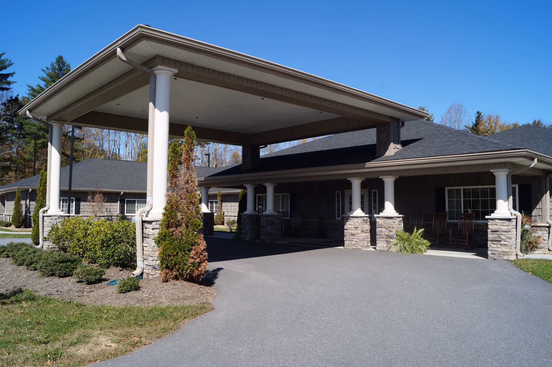 Exterior view of Mitchell House facility entrance with a covered driveway supported by white columns and stone bases, surrounded by shrubs and trees under a clear blue sky.