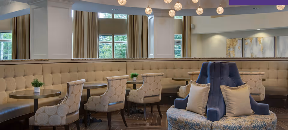 A cozy and elegant dining area with a curved beige tufted banquette along the wall, round tables with patterned beige chairs, small potted plants on the tables, large windows with beige curtains, and a central blue upholstered seating piece with beige pillows under hanging spherical pendant lights.