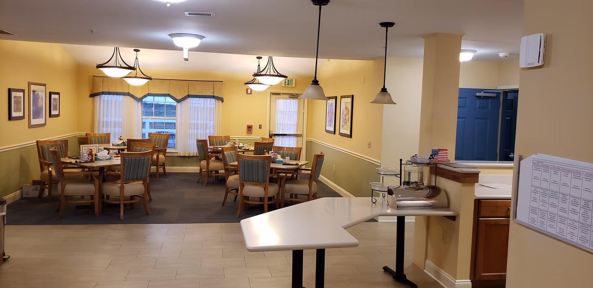 Interior view of a senior living community dining room with several round tables and wooden chairs with cushioned seats. The walls are painted yellow and green with framed artwork hanging. There are pendant lights hanging from the ceiling and large windows with curtains letting in natural light. A counter area with a small American flag and some kitchen items is visible on the right side.