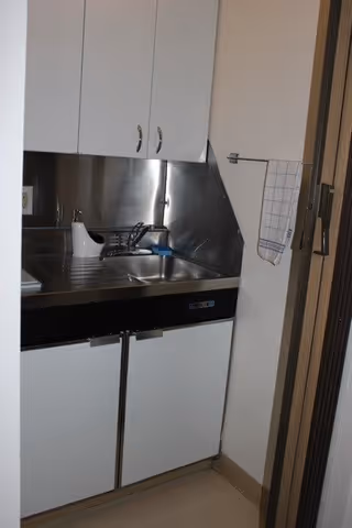 Small kitchenette area with a stainless steel sink, faucet, and countertop. White cabinets are mounted above and below the sink. A white soap dispenser and a blue sponge are on the countertop. A checkered dish towel hangs on a hook on the wall to the right.