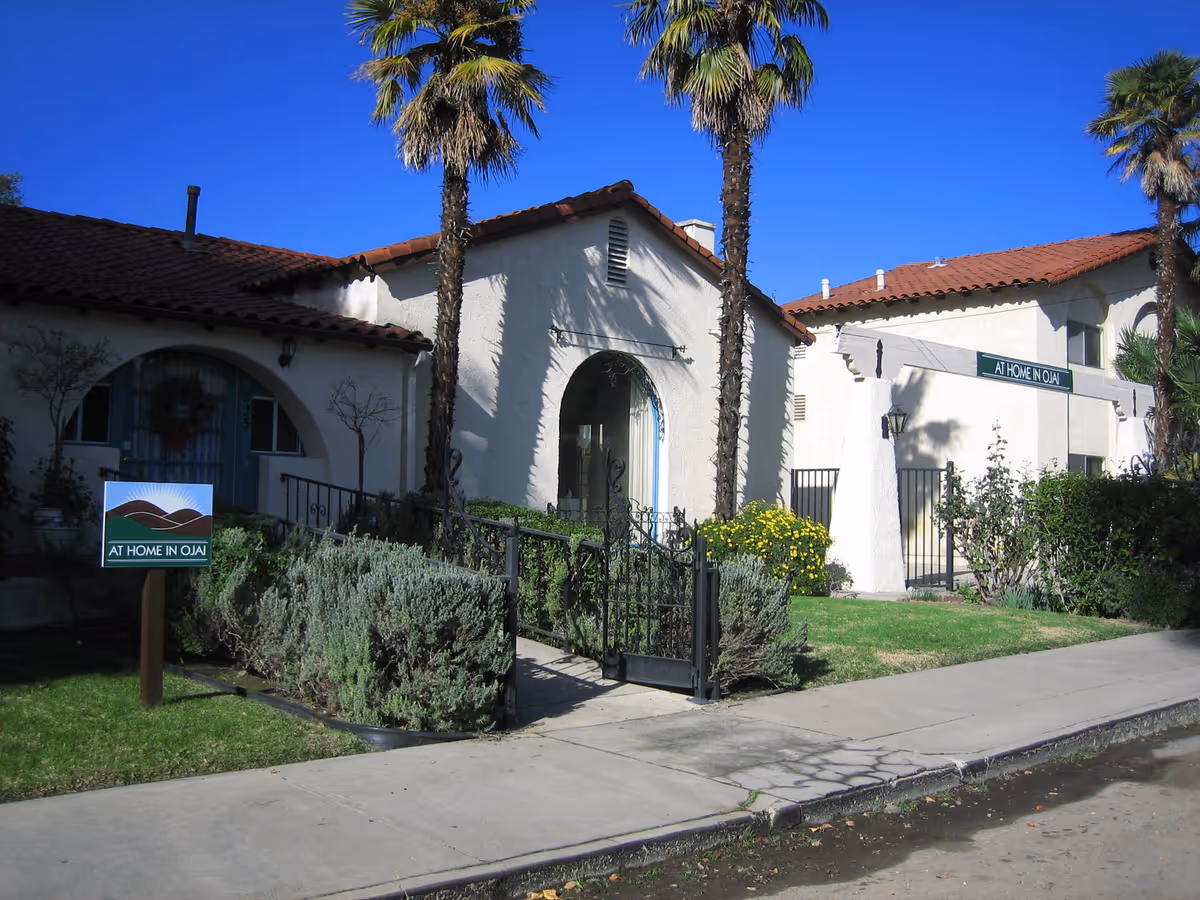 Front exterior of a Spanish-style building with palm trees, a gated walkway and a lawn sign that says "AT HOME IN OJAI".