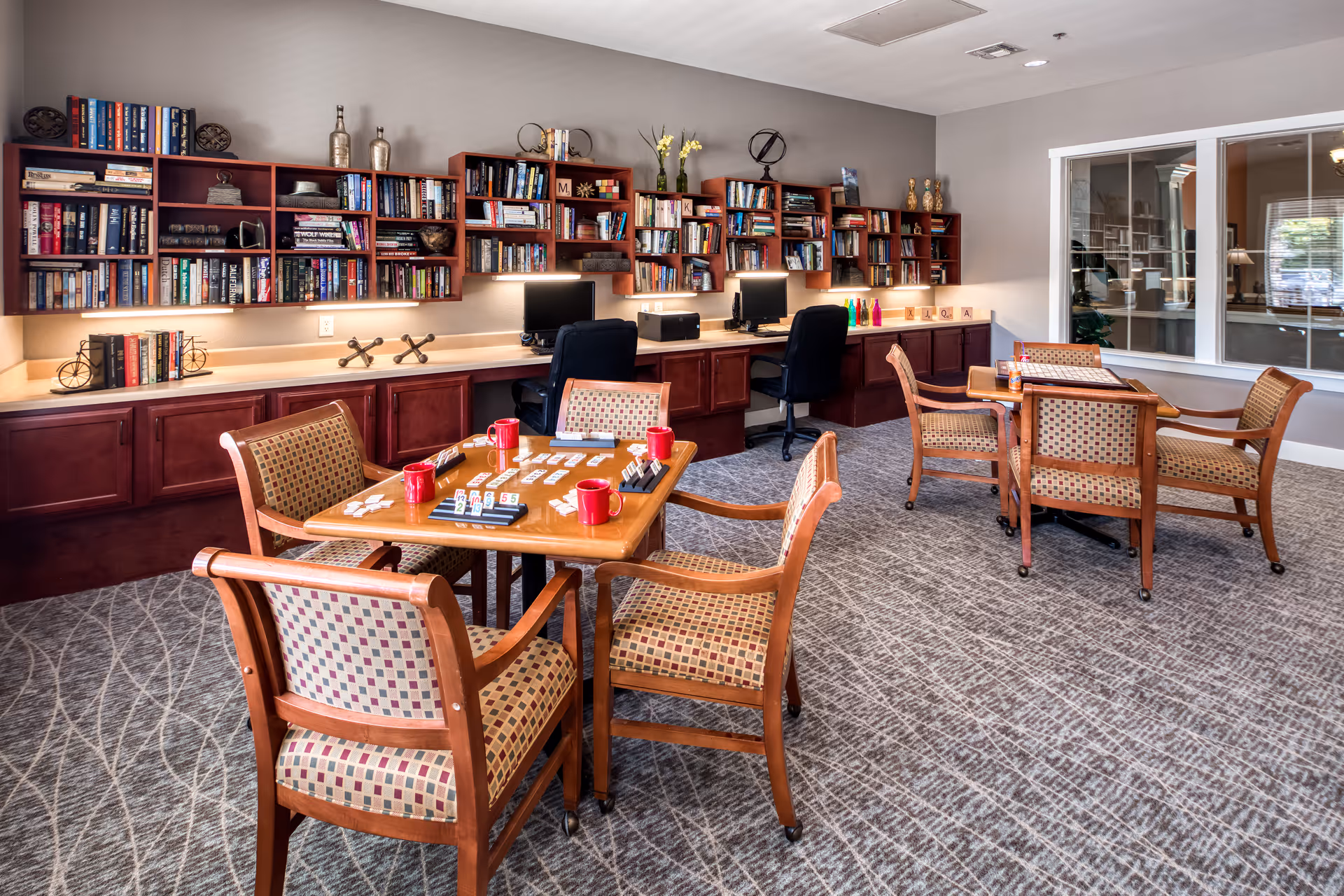 A cozy activity room in an assisted living facility featuring two wooden tables with patterned cushioned chairs. One table has a game set up with tiles and red mugs. The back wall has built-in wooden bookshelves filled with books and decorative items, with two computer workstations underneath. The room has a carpeted floor and a large window looking into another room.