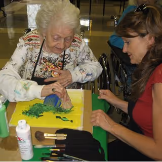 An elderly woman and a younger woman are seated at a table engaged in a painting activity. The elderly woman is using a sponge brush to apply green paint on a yellow canvas, while the younger woman watches and supports her. Various paintbrushes and a bottle of white paint are on the table.