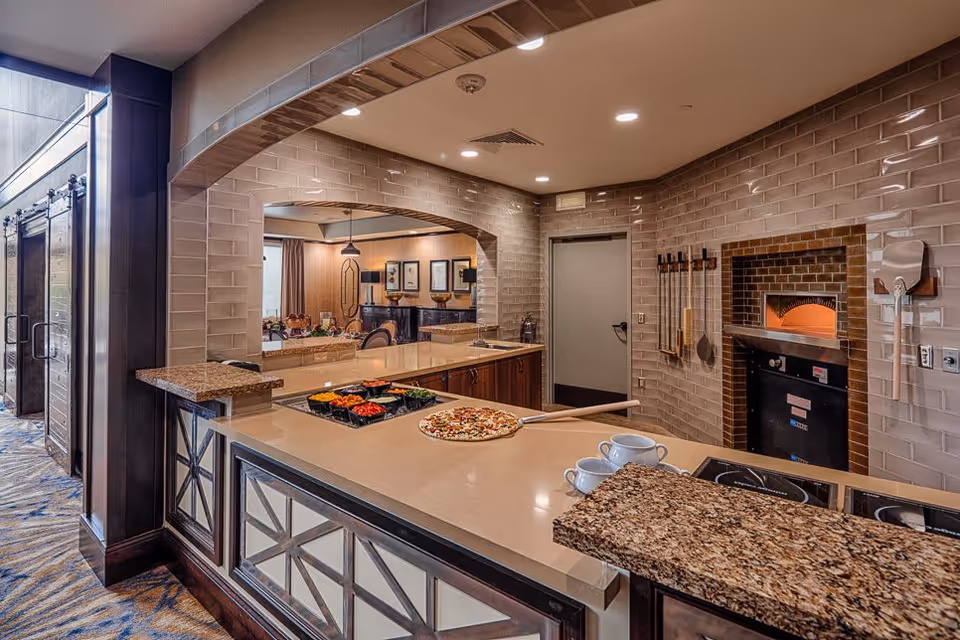 Interior view of a kitchen area in a senior living facility with a pizza oven built into a tiled wall. A pizza peel with a prepared pizza rests on the countertop, along with bowls of pizza toppings and two white cups. The kitchen has a granite countertop and an open window looking into a dining or common area with framed pictures on the wall.
