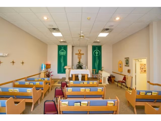 Interior view of a small chapel or prayer room with wooden pews arranged in rows facing an altar. The altar has a white cloth, a crucifix mounted on the wall behind it, and two green banners with religious symbols. There are hymnals or books placed on the pews, a statue of the Virgin Mary on the left side, and a piano on the right side near the altar.