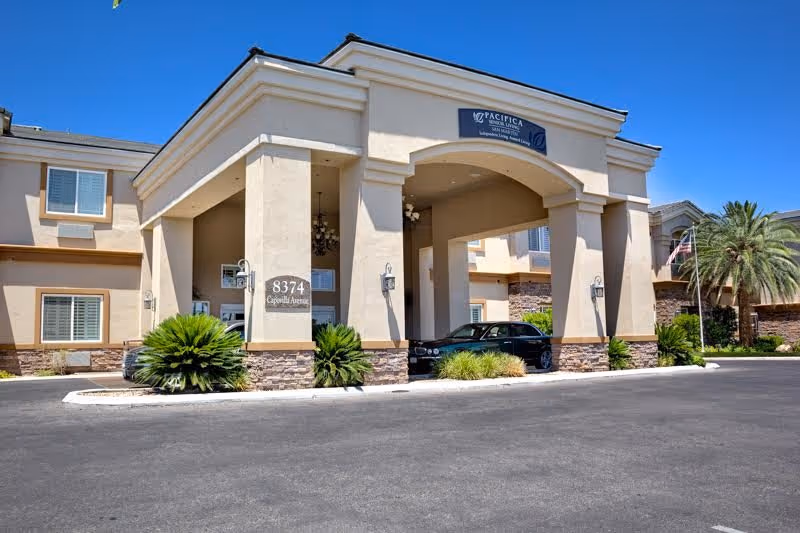 Exterior view of a senior living facility building with a covered entrance supported by columns. The building is beige with stone accents at the base and has several windows with white shutters. There are green plants and palm trees around the entrance, and a black car is parked under the covered area. The sky is clear and blue.