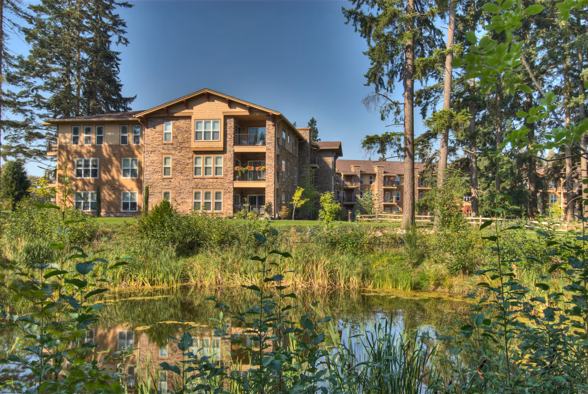 Exterior view of a multi-story senior living facility named Wesley Lea Hill surrounded by tall trees and greenery, with a small pond reflecting the building and vegetation in the foreground under a clear blue sky.
