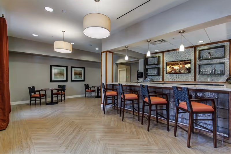 Communal dining and bar area with high stools at a long counter, pendant lights, and small table seating along a neutral wall.