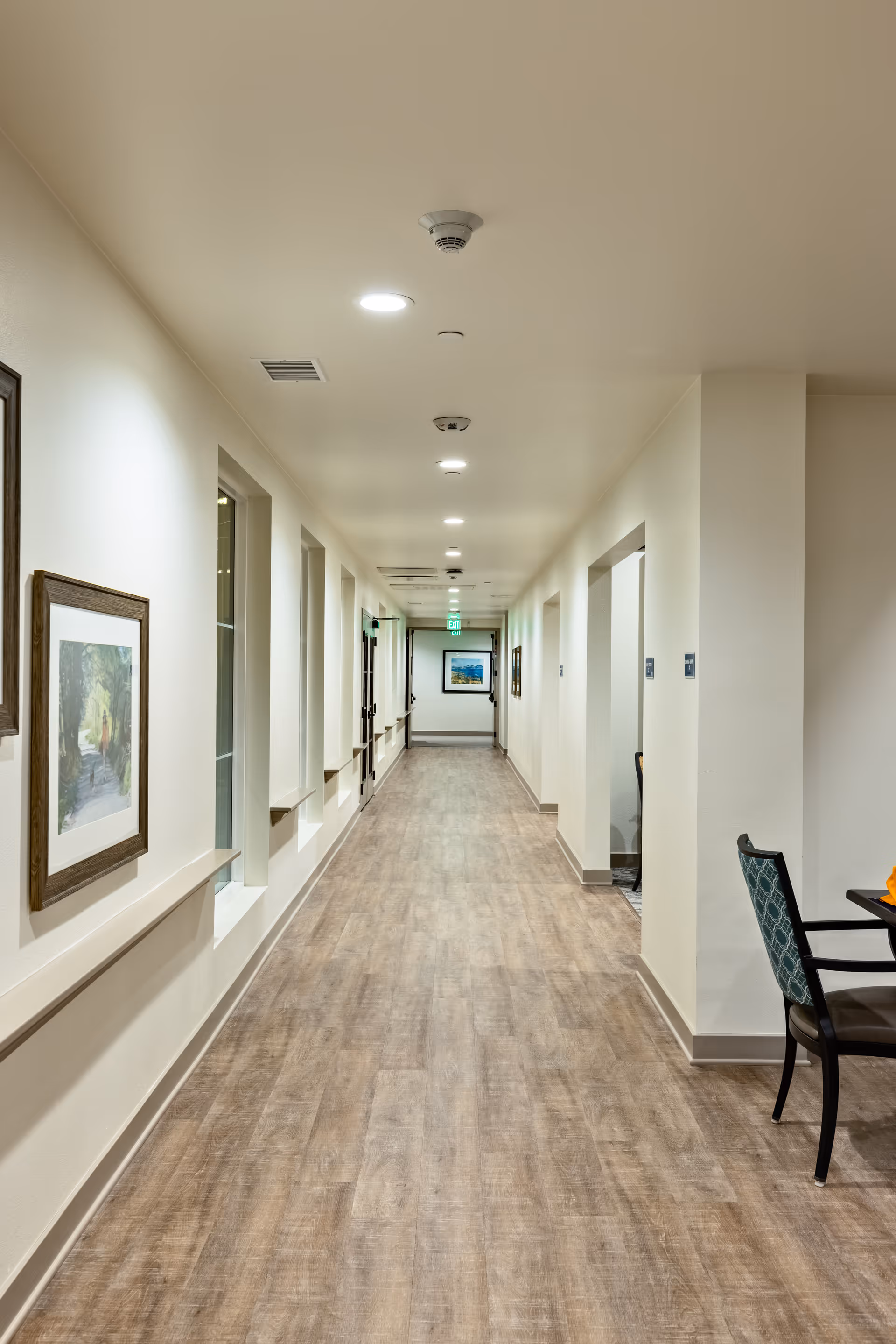 A long, well-lit hallway in a senior living facility with wood-look flooring, white walls, framed artwork on the left wall, and several doorways on the right. A chair and part of a table are visible on the right side near the foreground.