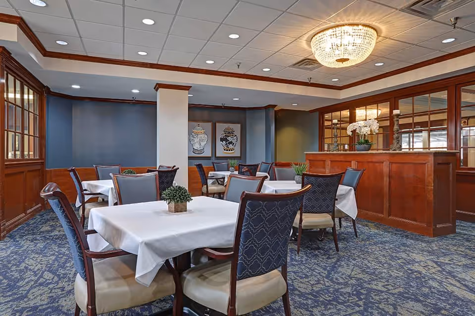 A dining room with several tables covered in white tablecloths, each surrounded by wooden chairs with cushioned seats and backs. The room features blue walls with wood paneling, a patterned carpet, framed artwork on the walls, and a large chandelier hanging from the ceiling. There is a wooden partition with decorative items and a mirror behind it.