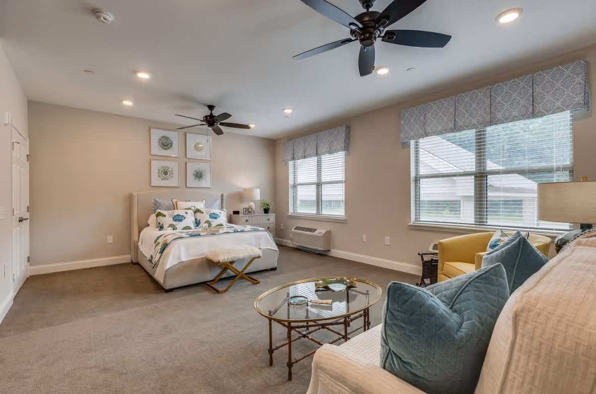 A spacious, well-lit bedroom with a bed adorned with floral bedding, a glass coffee table, a white sofa with blue pillows, a yellow armchair, two large windows with patterned valances, ceiling fans, and framed artwork on the wall.