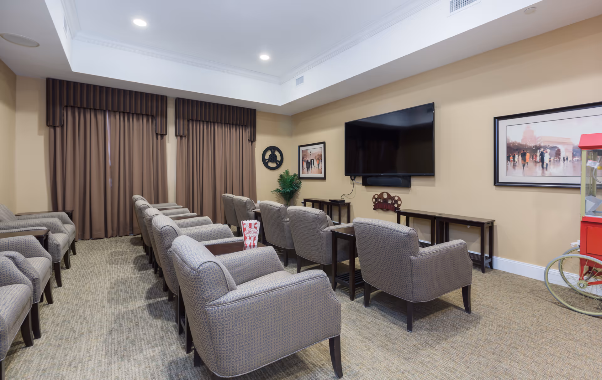 A cozy media room with two rows of comfortable armchairs facing a large flat-screen TV mounted on a beige wall. The room has brown curtains, framed artwork, a popcorn machine on the right, and a carpeted floor.