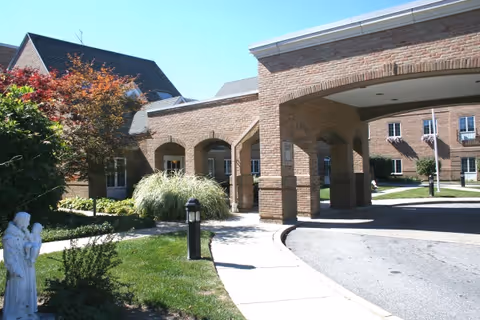 Front entrance of a brick senior living residence with a covered porte-cochere, walkway, and landscaped grounds.