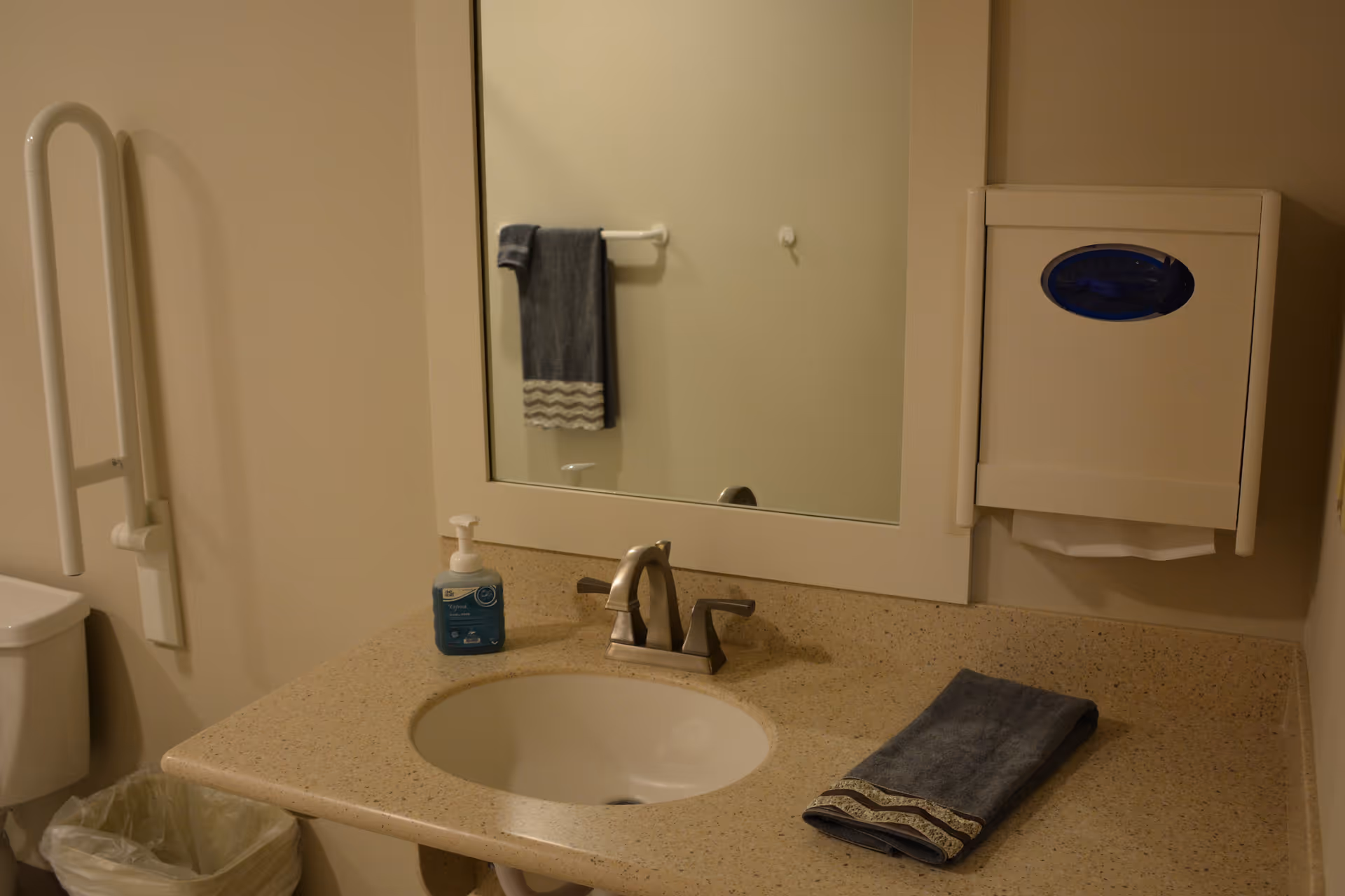 A bathroom sink area with a beige countertop, a silver faucet, a bottle of hand soap, and a folded dark gray towel with a decorative edge. A large mirror is mounted on the wall above the sink. To the right of the mirror is a wall-mounted paper towel dispenser. To the left, a grab bar is installed next to a toilet partially visible, with a trash can below.