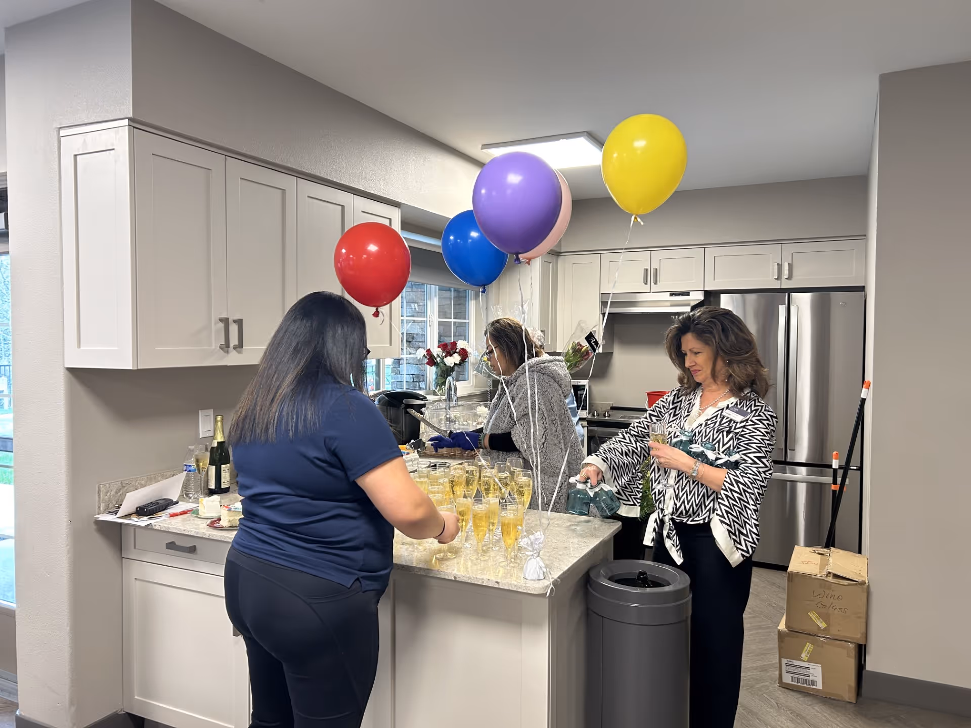 Three women in a modern kitchen preparing drinks on a countertop decorated with colorful balloons including red, blue, purple, pink, and yellow. The kitchen features white cabinets, a stainless steel refrigerator, and a trash can nearby. There are boxes stacked in the corner and a window letting in natural light.