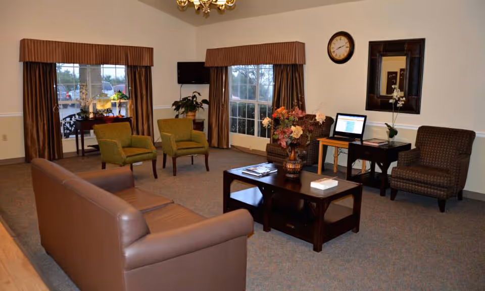 A cozy living room area with a brown leather sofa, two green armchairs, and two patterned armchairs arranged around a dark wooden coffee table with a floral centerpiece. The room has large windows with brown curtains, a wall clock, a wall mirror, a small table with a laptop, and a TV mounted on the wall.