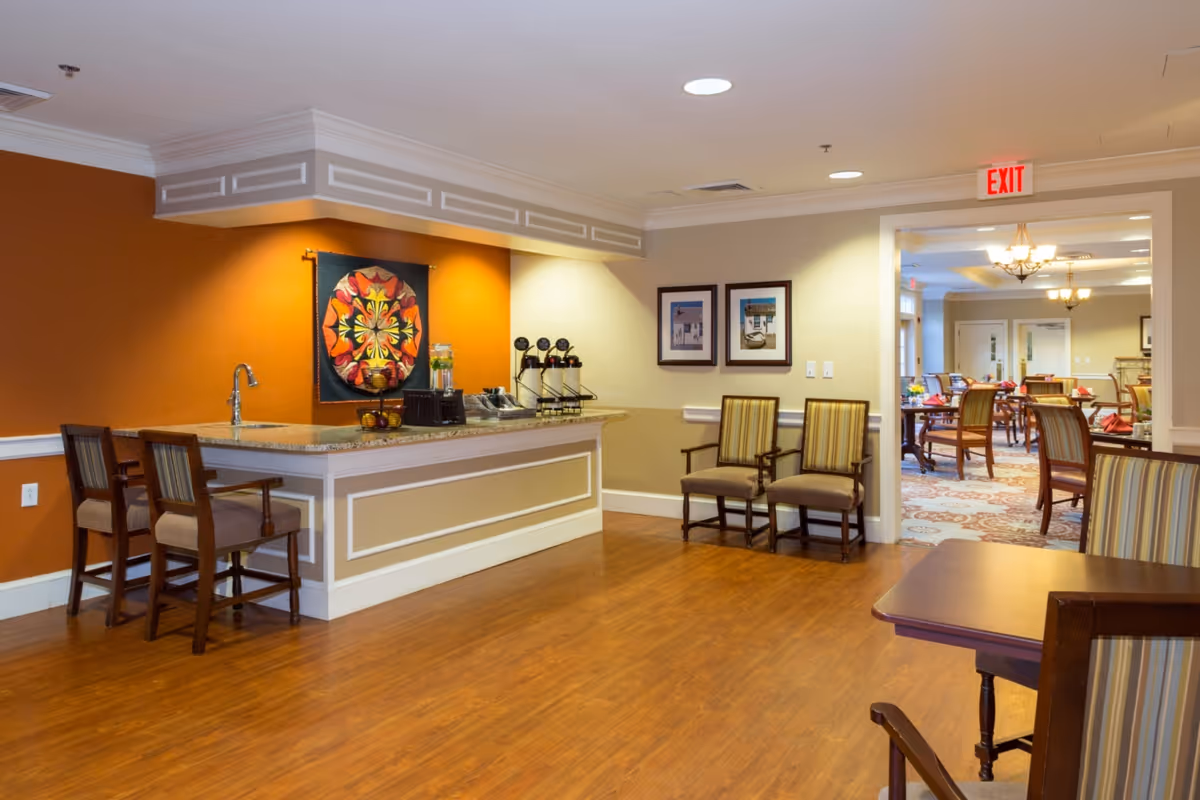Interior view of a senior living facility lounge area with a small counter featuring a sink, beverage dispensers, and fruit basket. There are two chairs at the counter and two additional chairs against the wall. The walls are painted in warm tones with framed pictures and a colorful quilt hanging on the wall. In the background, a dining area with tables and chairs is visible through an open doorway.
