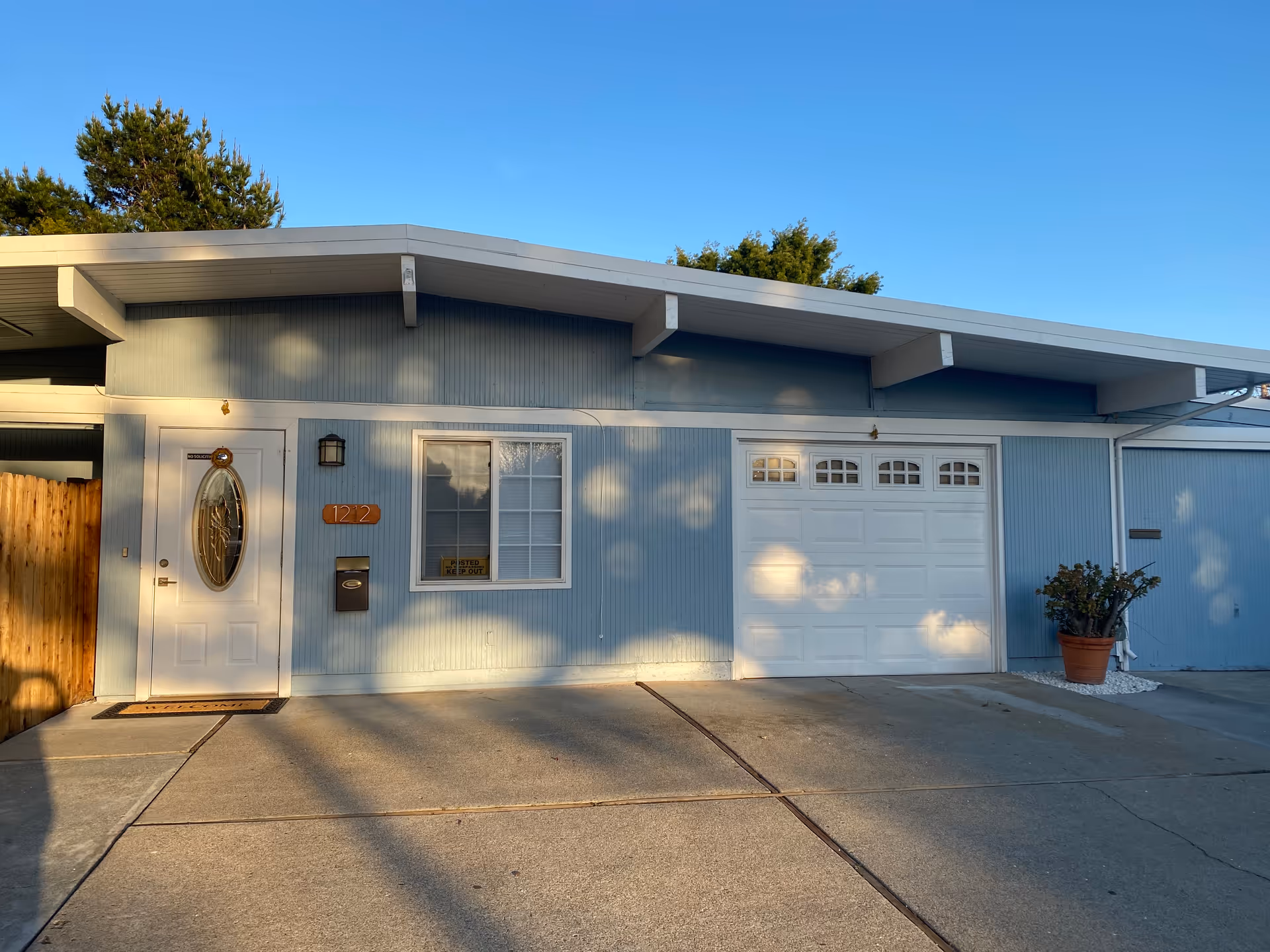 Light-blue single-story home's front facade with a white entry door, window, attached garage, and driveway under a clear sky.