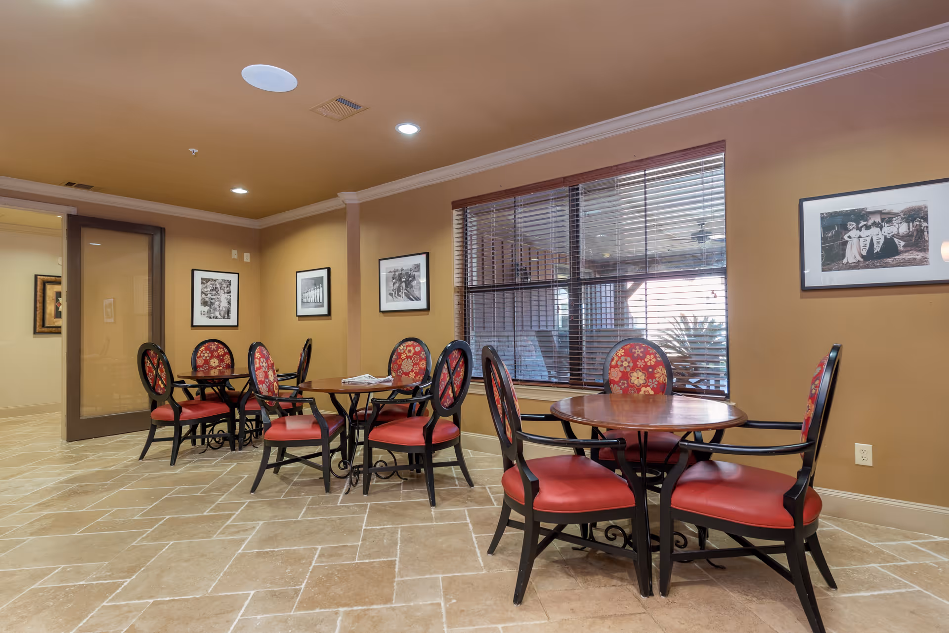 Interior view of a dining area with round wooden tables and red cushioned chairs with floral patterns. The room has beige walls, tiled flooring, and a large window with blinds. Black and white framed photographs hang on the walls.