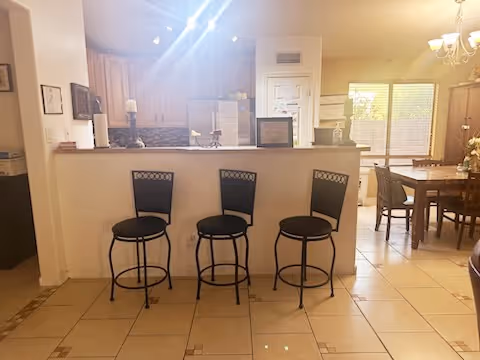 Interior view of a living space showing a kitchen counter with three black bar stools in front. Behind the counter is a kitchen area with wooden cabinets, a refrigerator, and a stove. To the right, there is a dining area with a wooden dining table and chairs near a window with blinds. The floor is tiled and the room is well-lit with natural light and ceiling lights.