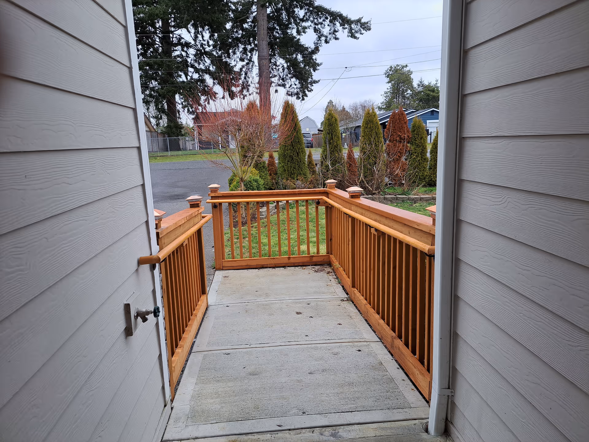 View through an open doorway onto a small concrete porch with wooden railings leading to a yard with shrubs and trees.