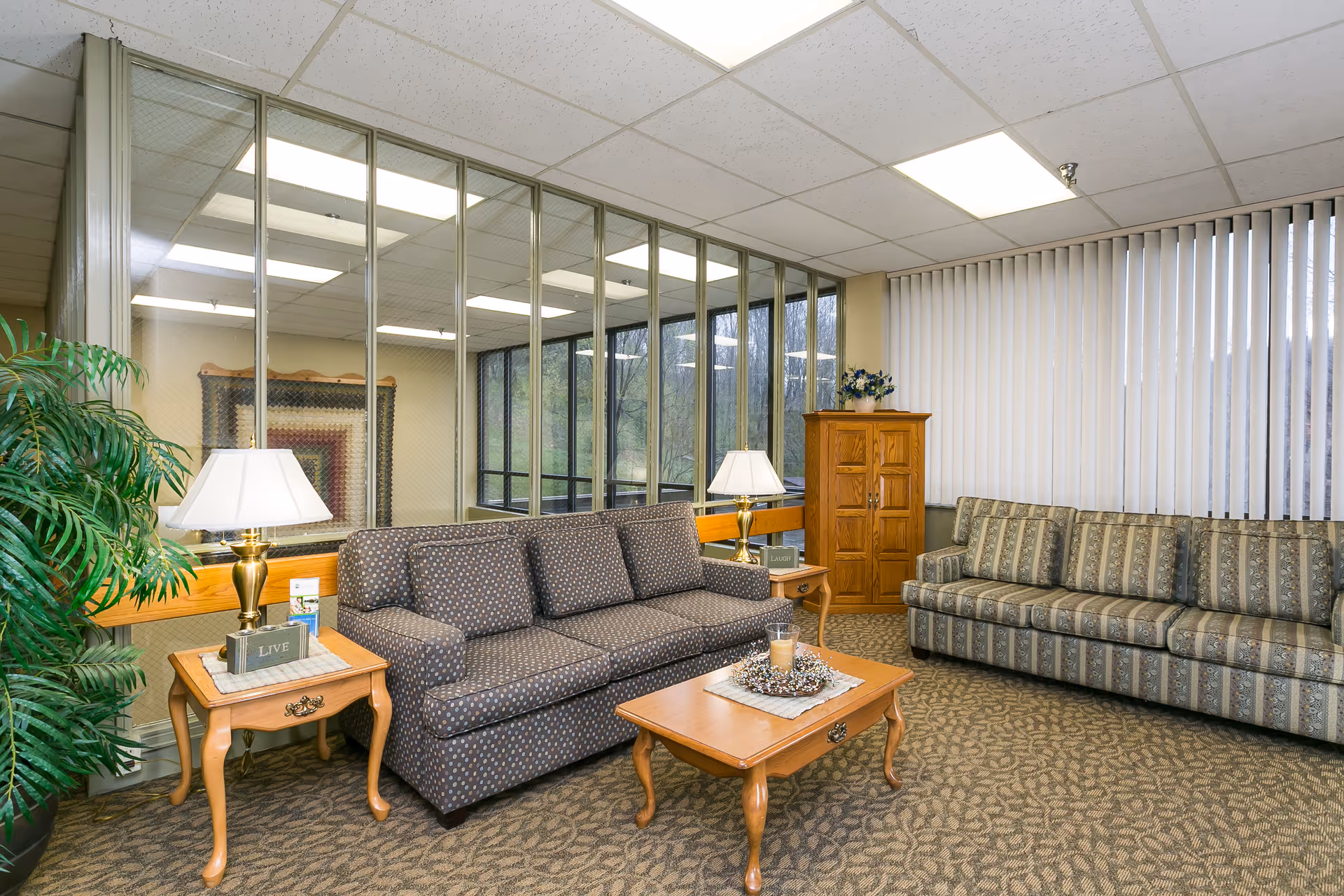 Carpeted sitting area with two patterned sofas, wooden coffee and side tables, lamps, a potted plant, and large windows with vertical blinds.