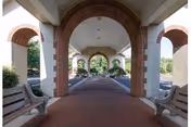 Covered walkway with brick arches and benches on either side, leading to an outdoor area with trees and greenery visible in the background.