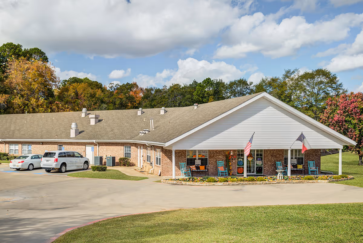 Exterior view of Winkler Court, a single-story brick building with a covered porch featuring rocking chairs and two flags. There are a few parked cars in front, surrounded by green grass and trees with autumn foliage under a partly cloudy sky.