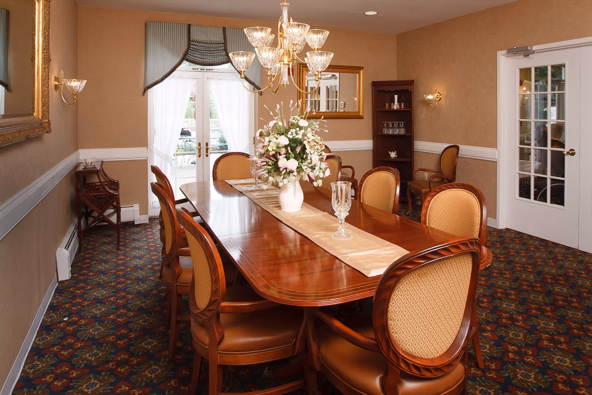 A formal dining room with a long polished wooden table surrounded by eight upholstered chairs. A floral centerpiece and crystal glasses are placed on a beige table runner. The room has patterned carpet, beige walls with white trim, a chandelier, wall sconces, a large mirror, a wooden shelving unit with glassware, and French doors with sheer curtains leading outside.