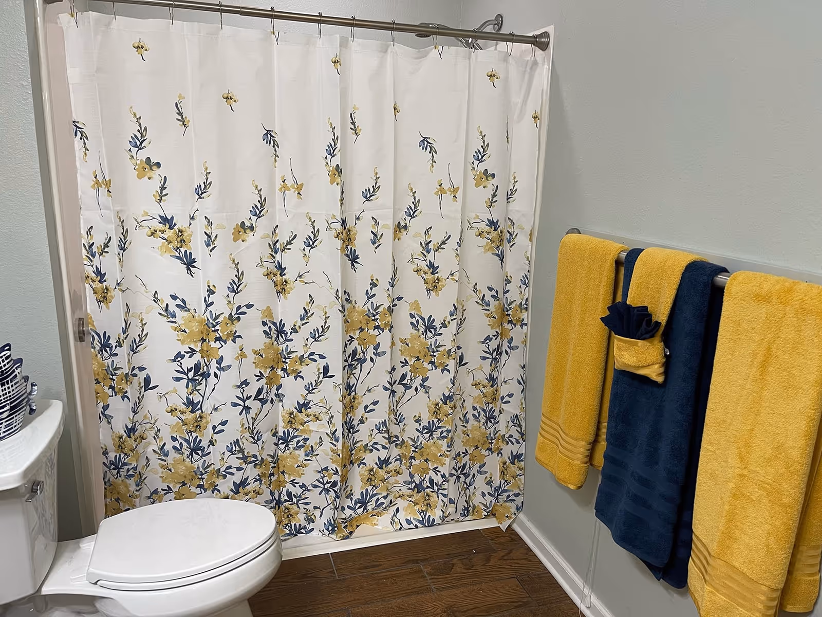 Bathroom with a white toilet, a shower with a white curtain featuring yellow and blue floral patterns, and a towel rack holding two yellow towels and one blue towel with a small rolled yellow and blue towel on top.