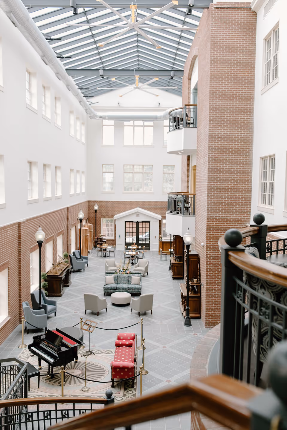 A spacious, bright indoor common area with high ceilings and large windows. The room features a black grand piano with a red cushioned bench, several seating areas with armchairs and sofas, and decorative streetlamp-style lights. The walls are a combination of white and exposed brick, and there are balconies overlooking the space. The floor is tiled with a geometric pattern.