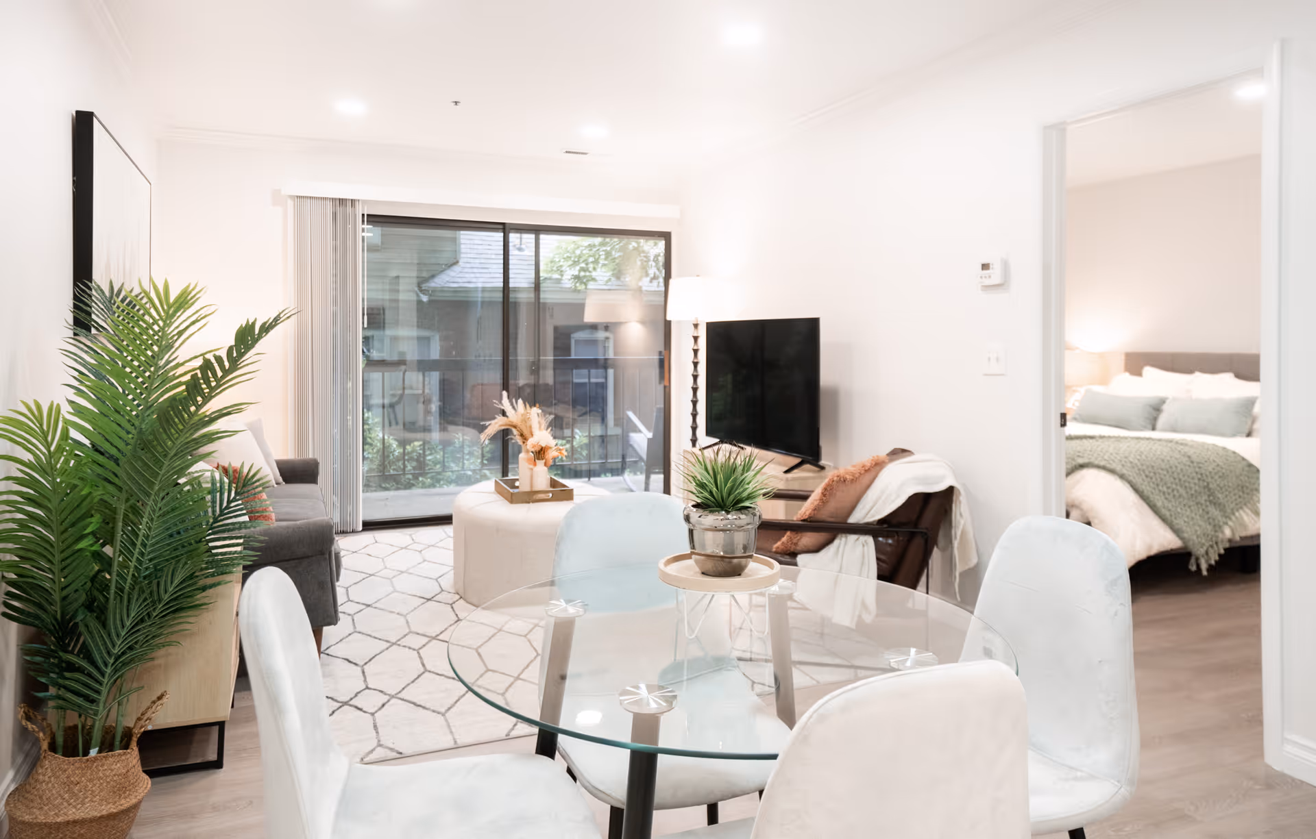 Bright modern living room and dining area with a glass table, potted plants, TV, and a view into a bedroom and sliding patio doors.