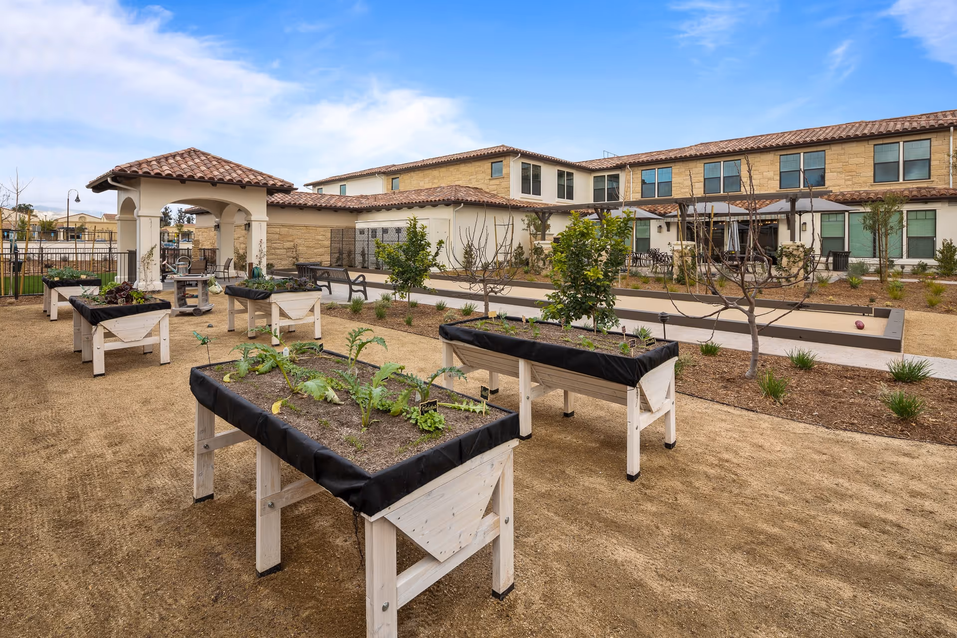 Raised gardening beds, benches, and a bocce court in an outdoor courtyard of a two-story senior living building under a blue sky.