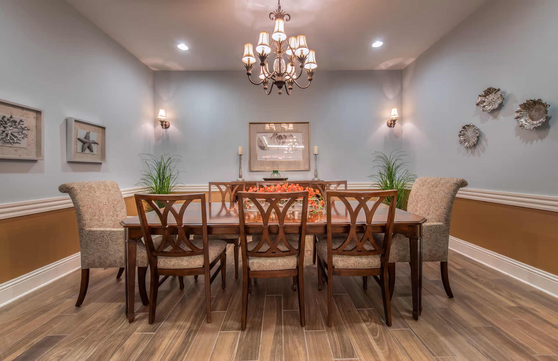 A formal dining room with a wooden dining table surrounded by six chairs, two upholstered armchairs at the ends and four wooden chairs with patterned cushions on the sides. The table has a centerpiece with red flowers. The room features light blue walls with wooden wainscoting, decorative wall art including framed starfish and metallic wall sculptures, two wall sconces with lampshades, and a chandelier hanging from the ceiling. The floor is wood with a natural finish.